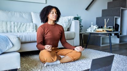 Woman sits cross legged with eyes closed in front of open laptop with a couch behind her