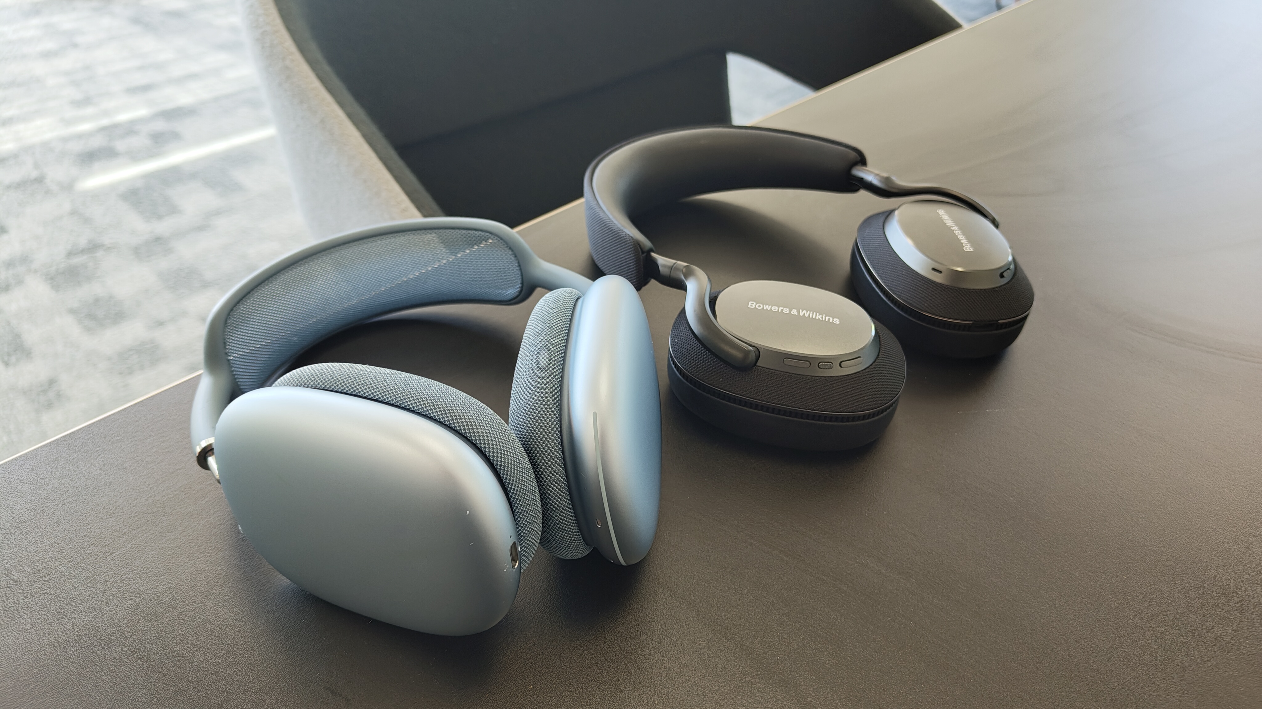 A blue pair of Apple AirPods Max headphone (left), next to a black pair of B&W Px7 S3 headphones on a desk.