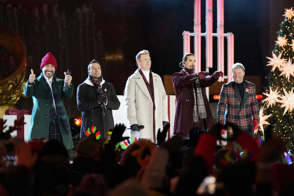 the backstreet boys wear coats and scarves standing next to a christmas tree while performing at the 2024 Christmas in Rockefeller Center special