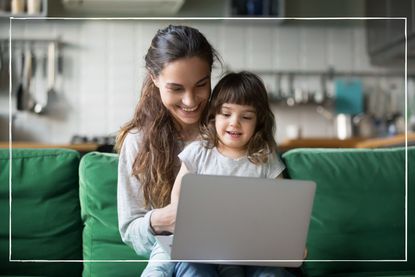 Mother and daughter smiling while sat together on a green sofa looking at laptop