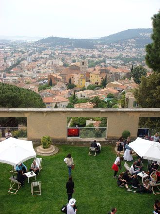 View of Hyères from the Villa Noailles. A green lawn with tables, chairs, people and a view of the city below.