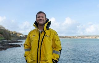 Dermot O'Leary wearing an RNLI branded yellow jacket, standing on the shore