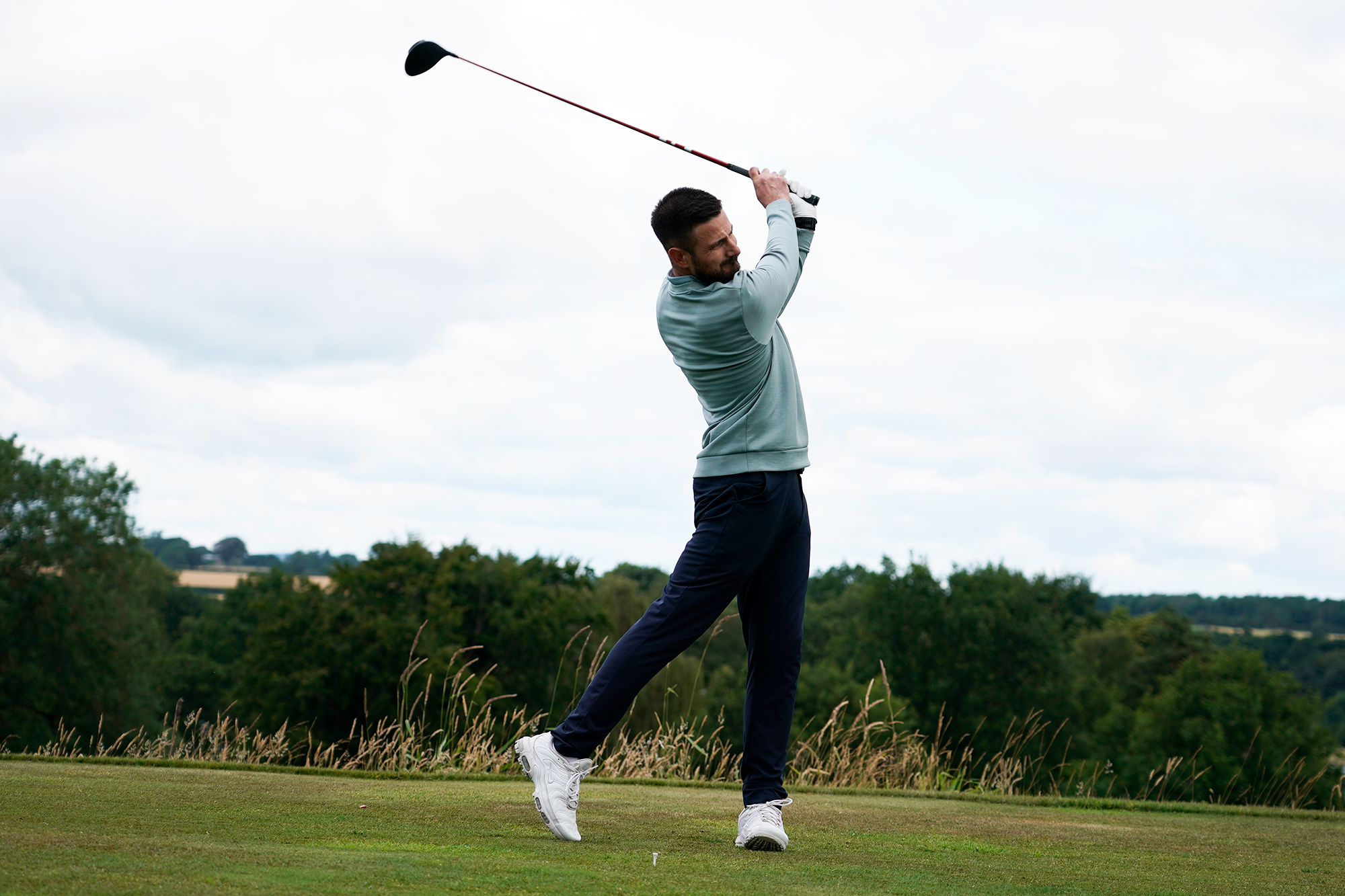 Baz Plummer hitting a driver, rising up into the finish position against a backdrop of the 16th hole at Sand Moor Golf Club