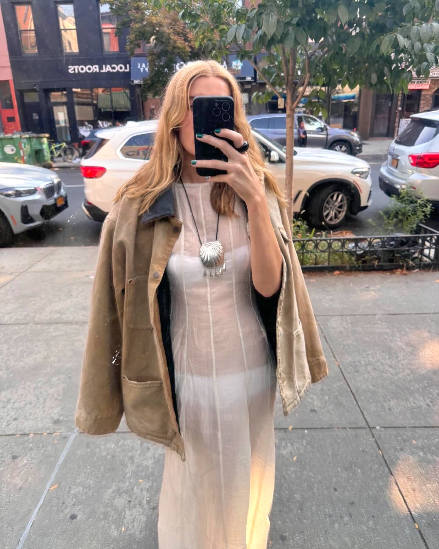 NYC-based style influencer Courtney Grow poses for a window reflection selfie wearing a canvas barn jacket, sheer white dress, and a silver Juju Vera Petra shell necklace