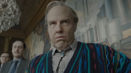 Tom Cruise is shown as Digger Rockwell, seated at a conference table wearing a multi-colored striped sweater and bolo tie, looking nervous in a screenshot from the Digger trailer.