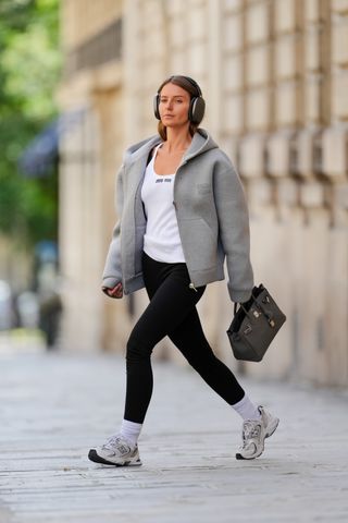 Diane Batoukina wears Apple airpods Max silver metallic headphones, a white Miu Miu t-shirt, a gray oversize Miu Miu hoodie sweater, black leggings from Zara, a Hermes Birkin bag, white socks, New Balance sneakers shoes, during a street style fashion photo session, on May 16, 2024 in Paris, France.