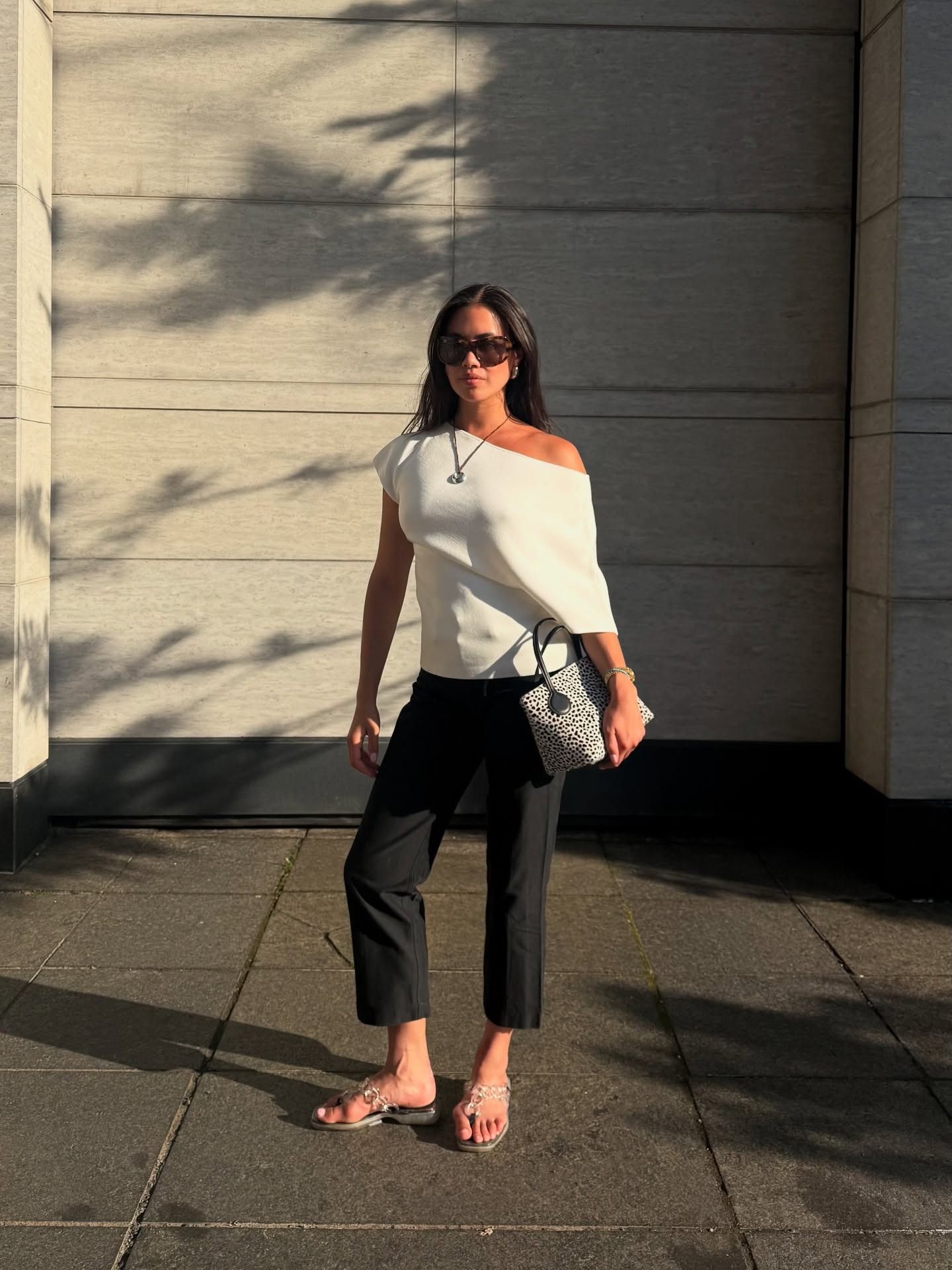Woman wears black cigarette pants, a Rohe structured top, clear flip flops and dalmation print bag.