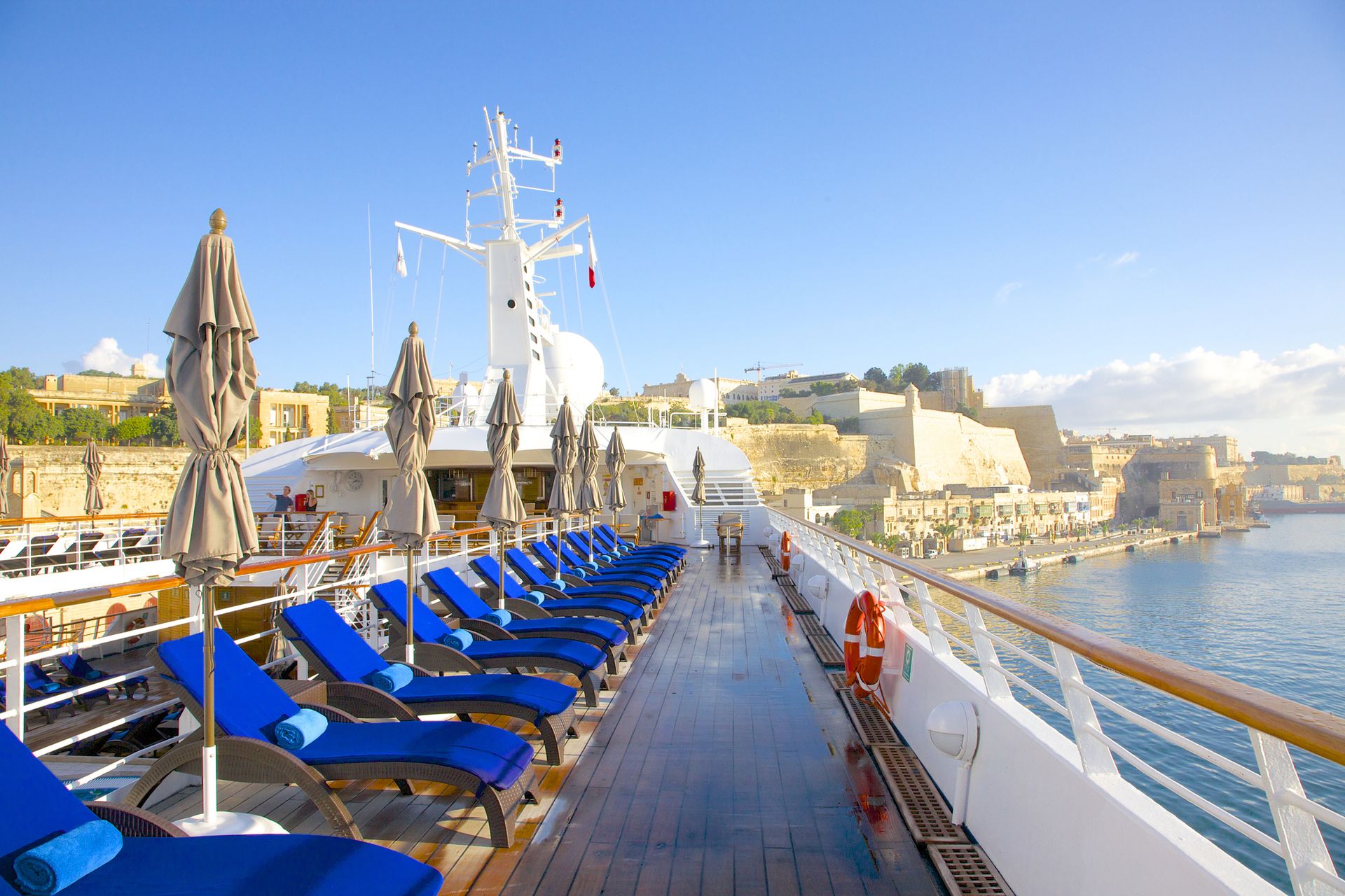 Early morning sunshine on teak deck and royal blue cushions awaiting passengers on Windstar yacht Star Legend. Yacht is docked near ancient fortification walls in Valletta, Malta.