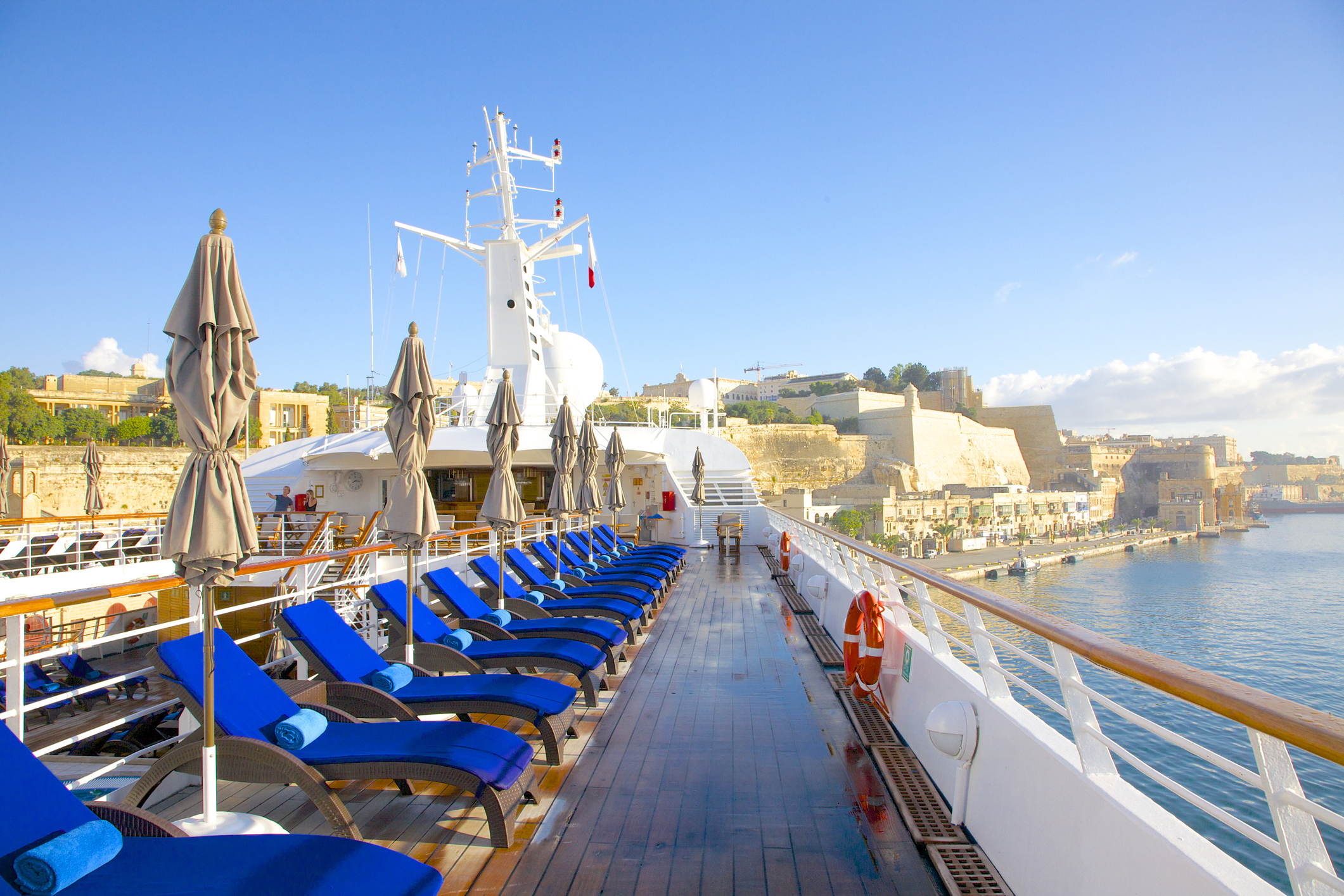 Early morning sunshine on teak deck and royal blue cushions awaiting passengers on Windstar yacht Star Legend. Yacht is docked near ancient fortification walls in Valletta, Malta.