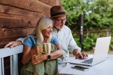 An older couple sits on a bench outside their rustic home, looking at a laptop.