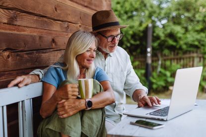 An older couple sits on a bench outside their rustic home, looking at a laptop.