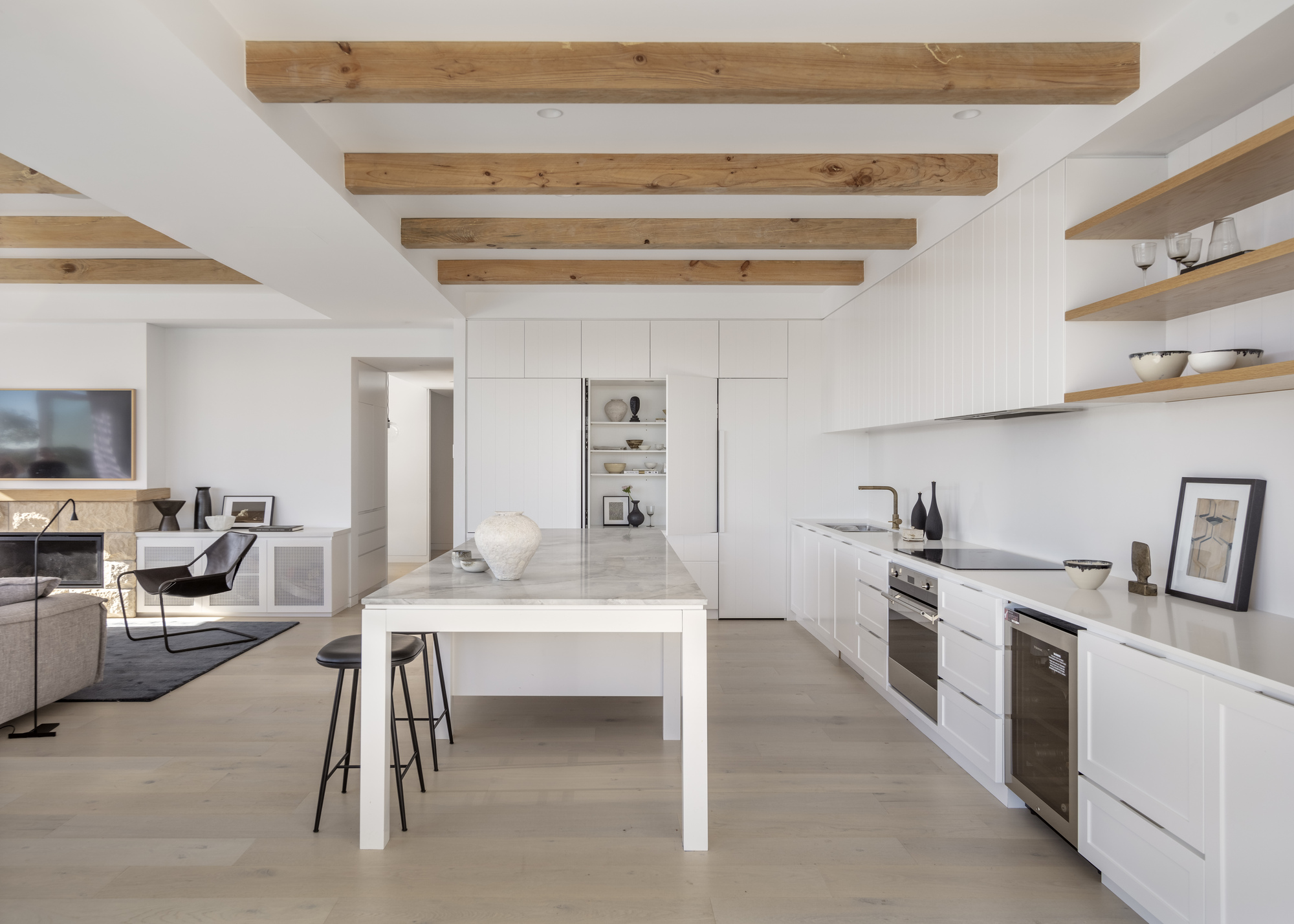 An open plan kitchen with white cabinets, wooden beams, and an adjoining living area
