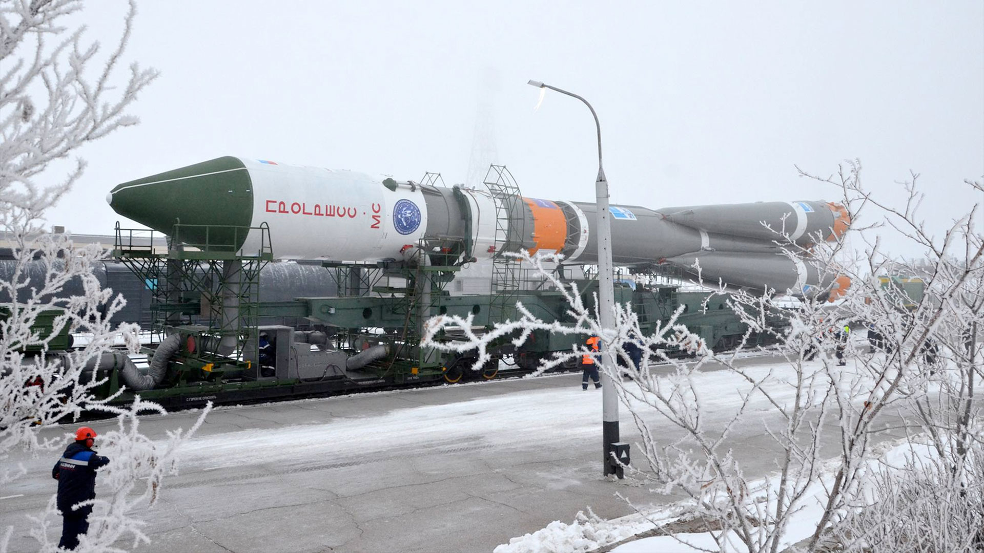 a greenish-gray, orange and white rocket is transported by rail to its launch pad among a snowy landscape