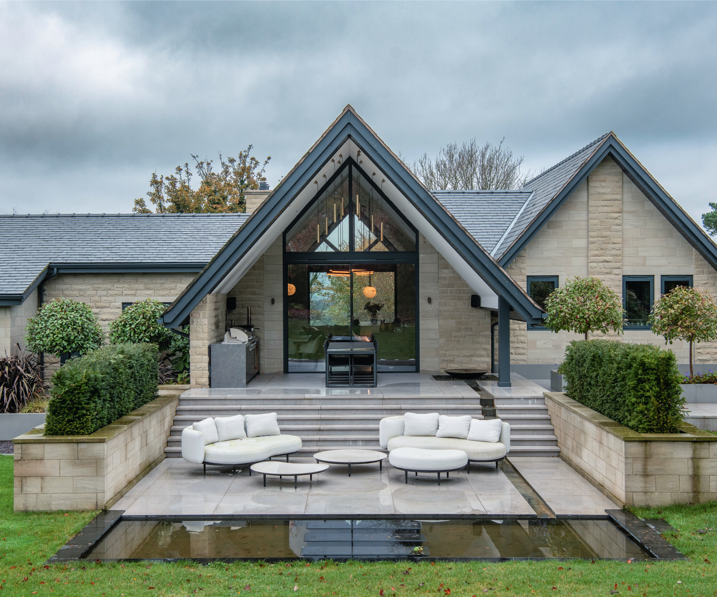 modern cream stone clad house with pitched gable, overhang and large gable end window and patio doors leading to terrace, floor glazing in garden showing basement below
