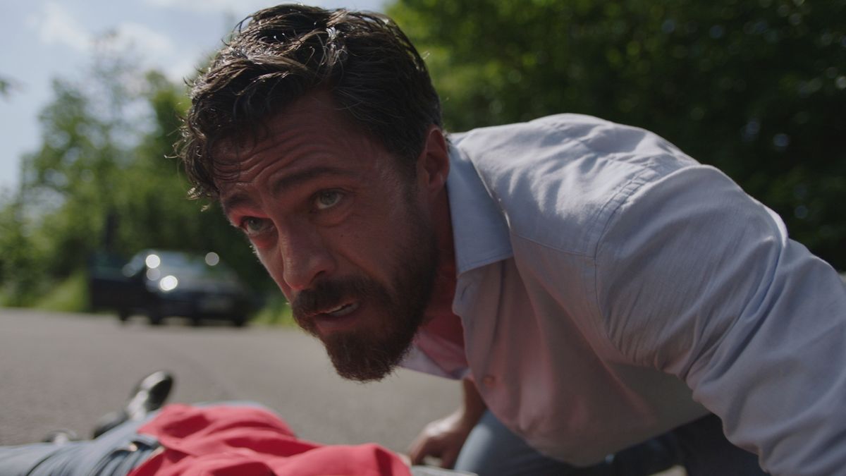 Olly Rix as Flynn Bryon. Close up shot of Flynn checking on an unconscious woman in a road. 