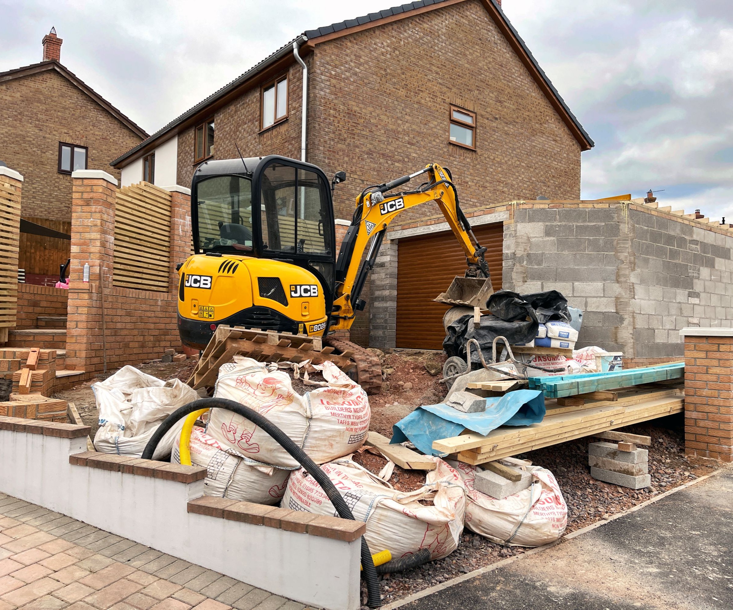 A yellow digger with a breeze block half built garage and a semi detached house behind
