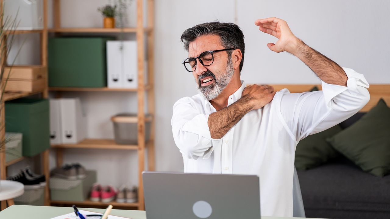 Mature man stretching and massaging shoulder at desk