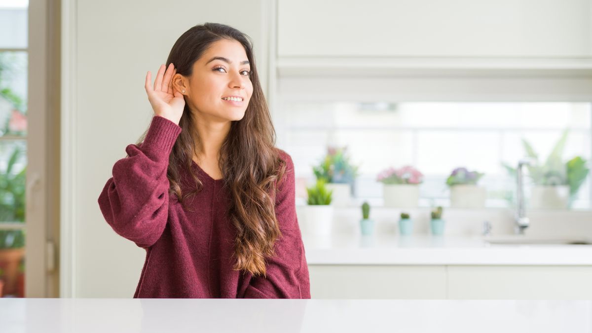 Young Asian woman sat at kitchen island with long dark hair, wearing red jumper with hand raised to ear as if listening