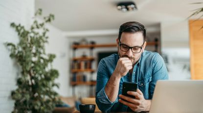 Pensive man looking at smart phone, sitting at table at home.