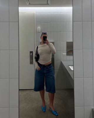 A woman taking a mirror selfie wearing long denim shorts.