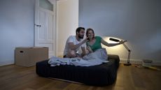 A couple sit on a mattress covered in black sheets in a new bedroom they've just moved into