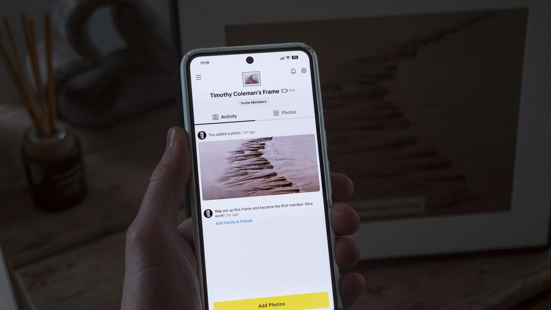 A phone being held, displaying the Aura companion app for digital photo frames, in front of the The Aura Ink 'E Ink' digital photo frame on a sideboard