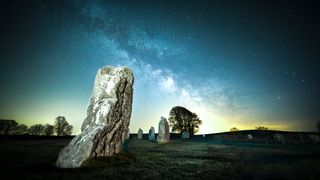 a large stone from a stone circle stands in the foreground. other stones dot the background, along with a large tree. light pollution shines in the sky along with the milky way