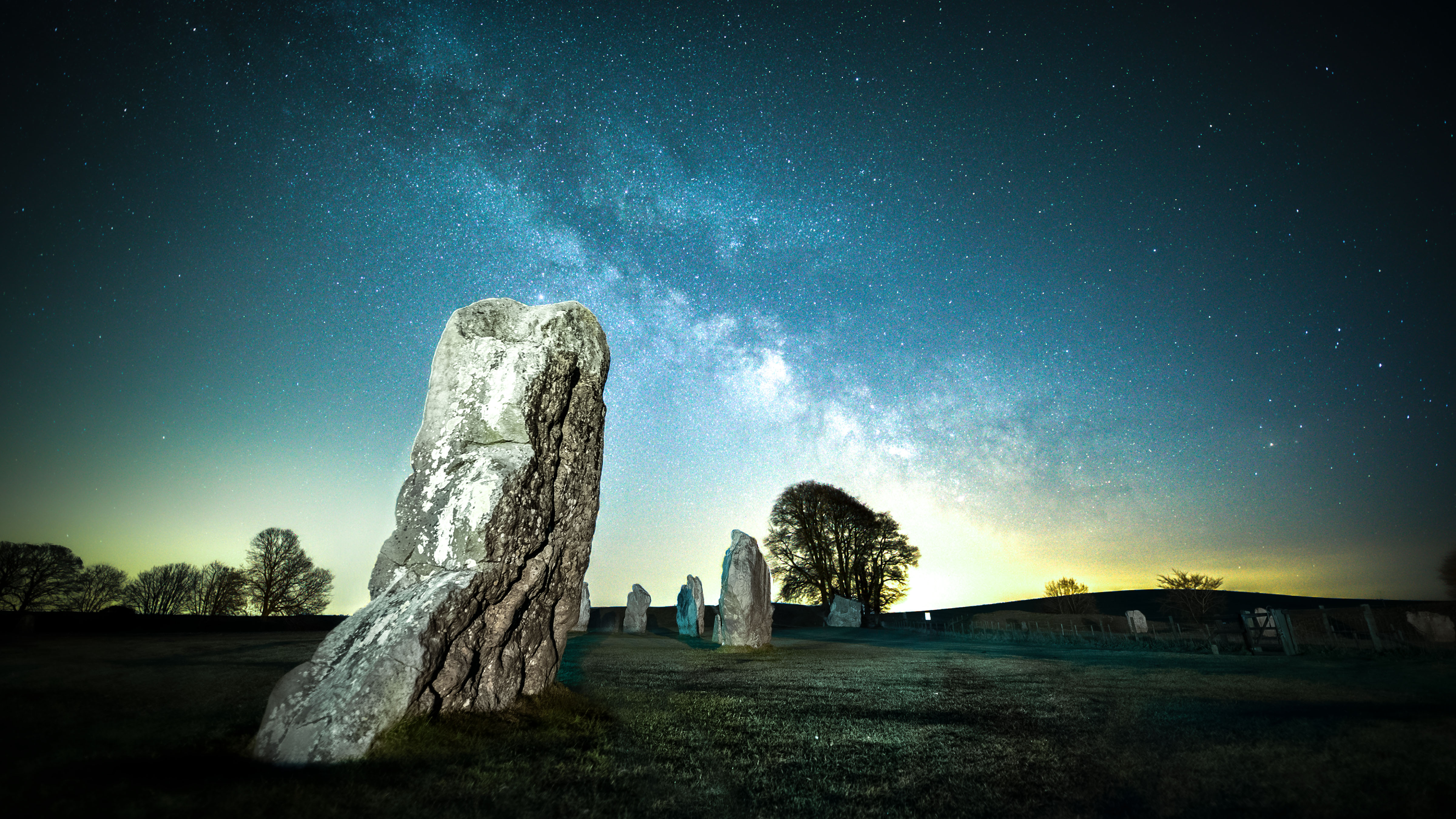 a large stone from a stone circle stands in the foreground. other stones dot the background, along with a large tree. light pollution shines in the sky along with the milky way