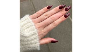 A close-up of Digital Beauty Writer, Sennen Prickett's long dark mauve almond nails/ with a pavement background