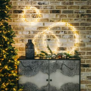 a sideboard beside a lit christmas tree with decorations on top and three lit circular garlands hanging on a brick wall behind