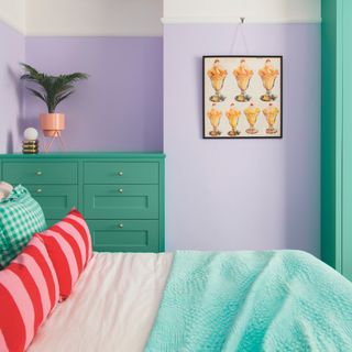tiny guest room ideas, colourful bedroom with colour blocking effect, lilac walls, green chest of drawers, artwork, red stripe cushions on bed, green throw