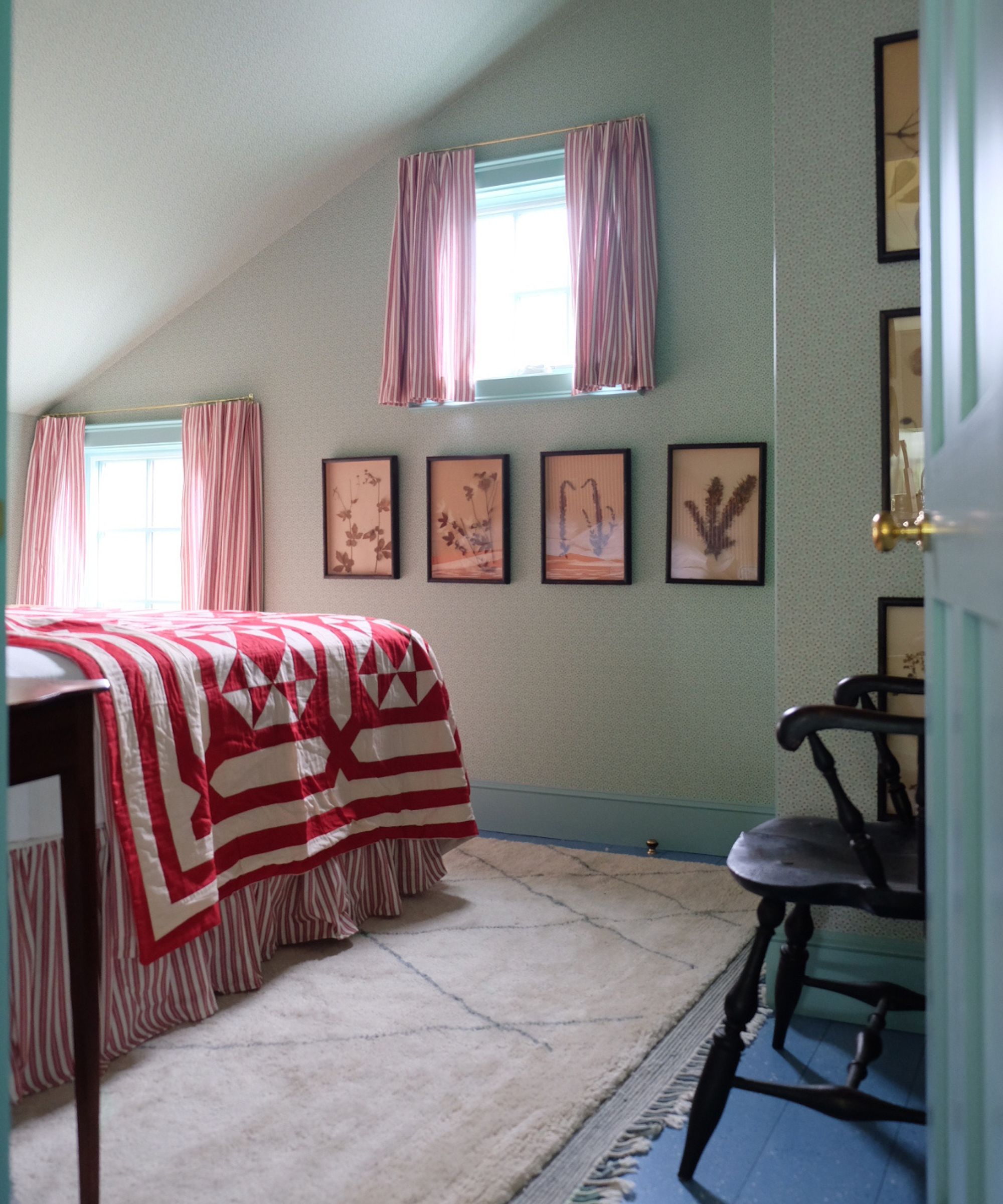 a vaulted blue bedroom with small floral wallpaper, blue painted floorboards, small windows with red striped curtains and a bed made with a red striped bed skirt and patchwork blanket