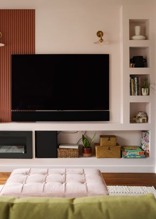 A living room with a tv on the wall. On the right side of the tv is narrow open shelving with books and other household essentials. Under the tv there is extra storage