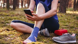 Injured hiker with sprained ankle and running first aid kit using smart phone to get help