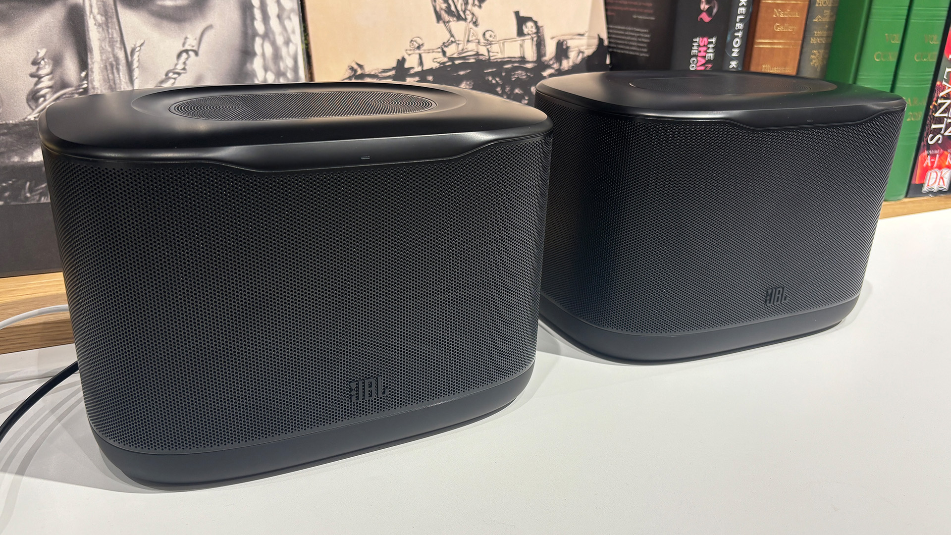JBL Cove X1 multi-room wireless speakers on white table in front of busy bookshelf