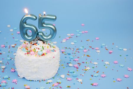 Coconut birthday cake on blue background with burning candles number 65 and many colorful sprinkles.