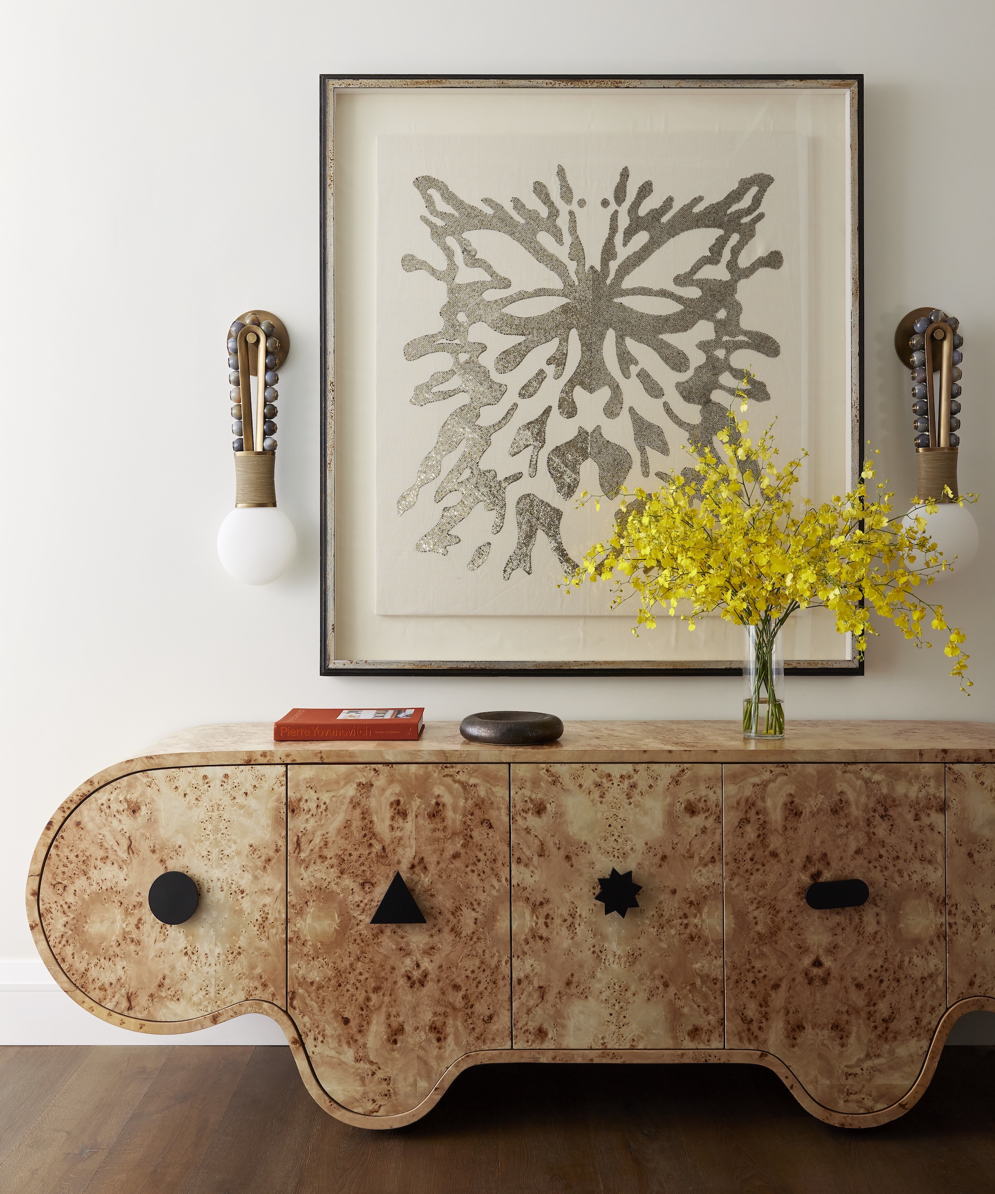 Curved sideboard with large artwork and wall lights above, and vase with yellow flowers, book and vessel displayed on it