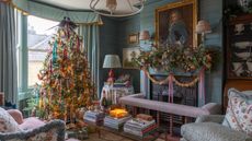 traditional english country living room wallpapered in blue grasscloth styled for christmas with a large velvet ribbon adorned tree topped with two faux pheasants and a fireplace styled with a large garland and dried oranges