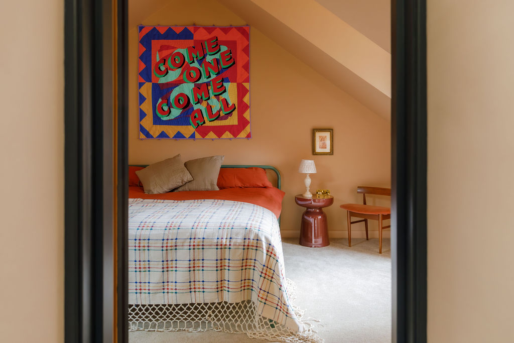 A bedroom with caramel-tinted walls, velvety gray carpet, orange bedsheets topped by a checkered white, orange, and blue blanket, a colorful quilt reading "come on, come all", and sculptural pieces.