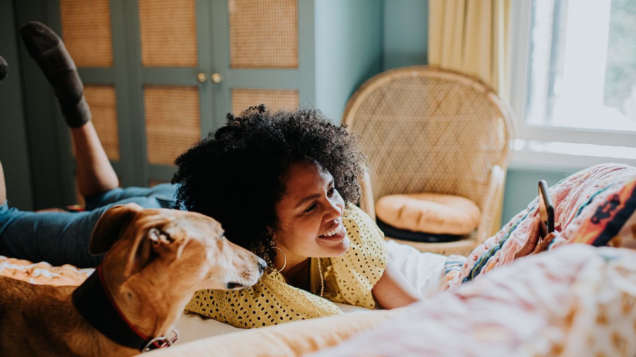 A relaxed-looking young woman hangs out on her bed with her dog, a smartphone in her hand.