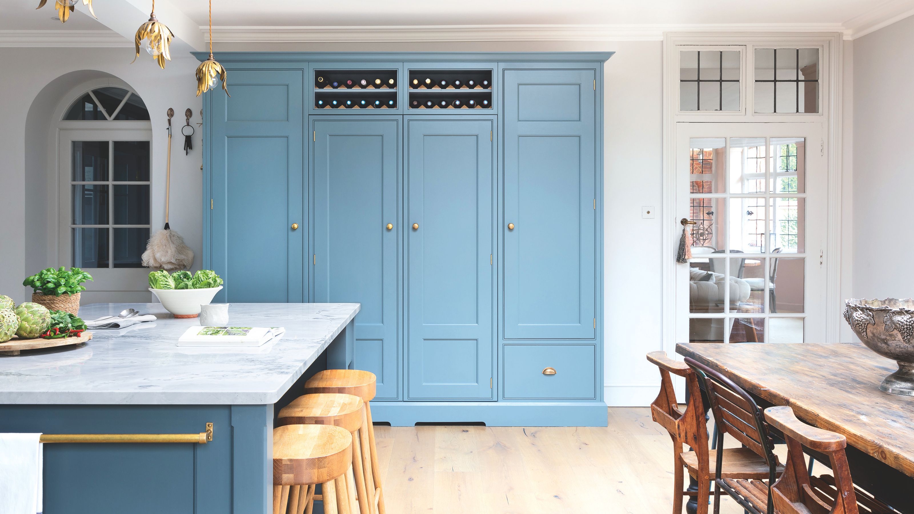 White painted kitchen-diner with pastel blue cabinets, a wooden dining room table on the right and a blue island on the left with a marble worktop
