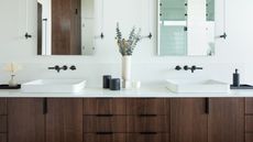 A double sink bathroom vanity with a vase in the middle and a leafy green plant inside as well as other bathroom accessories on top of the counter. There is also a view of double mirrors with lights on each side