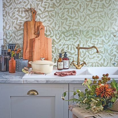 Kitchen sink with lots of cooking accessories next to it, brass taps and cupboard handles, and green foliage wallpaper behind it