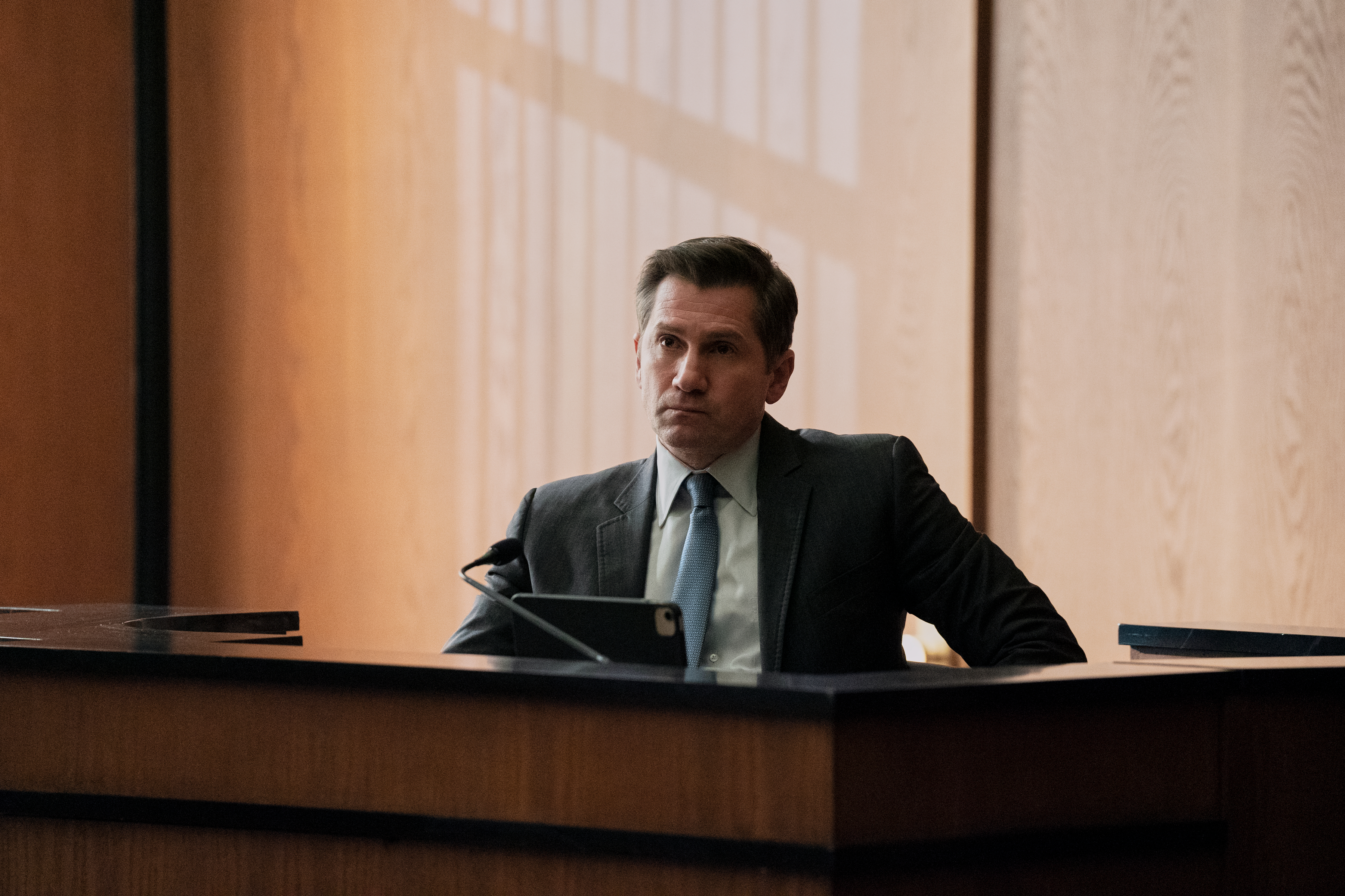 A man wearing a suit (Michael Goorjian as Alex Grant) sits in the witness box in a courtroom, in episode 209 of 'The Lincoln Lawyer.'