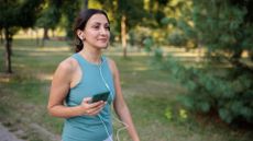 Woman walking with headphones in and holding phone in outdoors next to trees, smiling