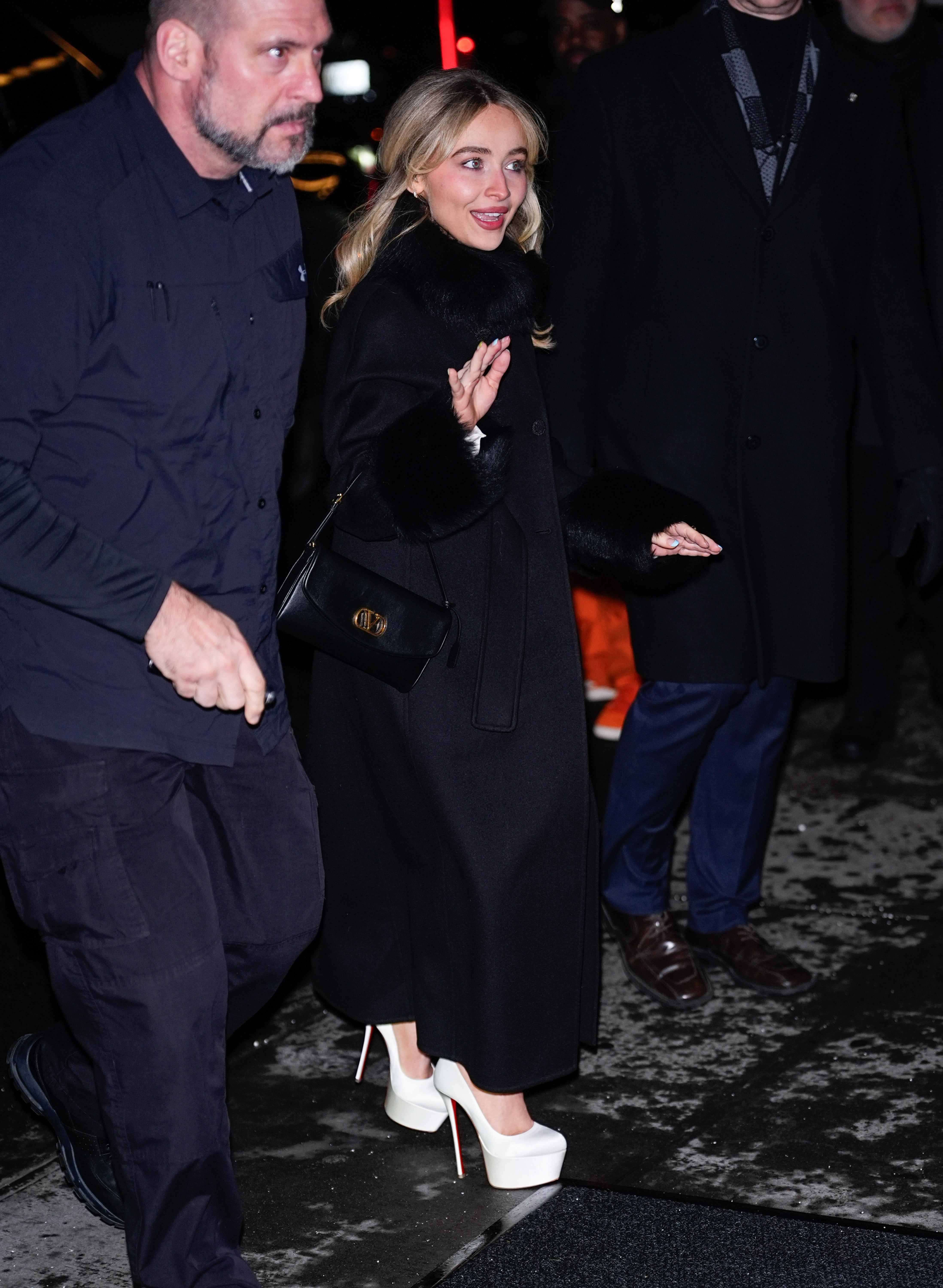 Sabrina Carpenter wears white satin platform heels with a longline black coat and a black leather handbag.
