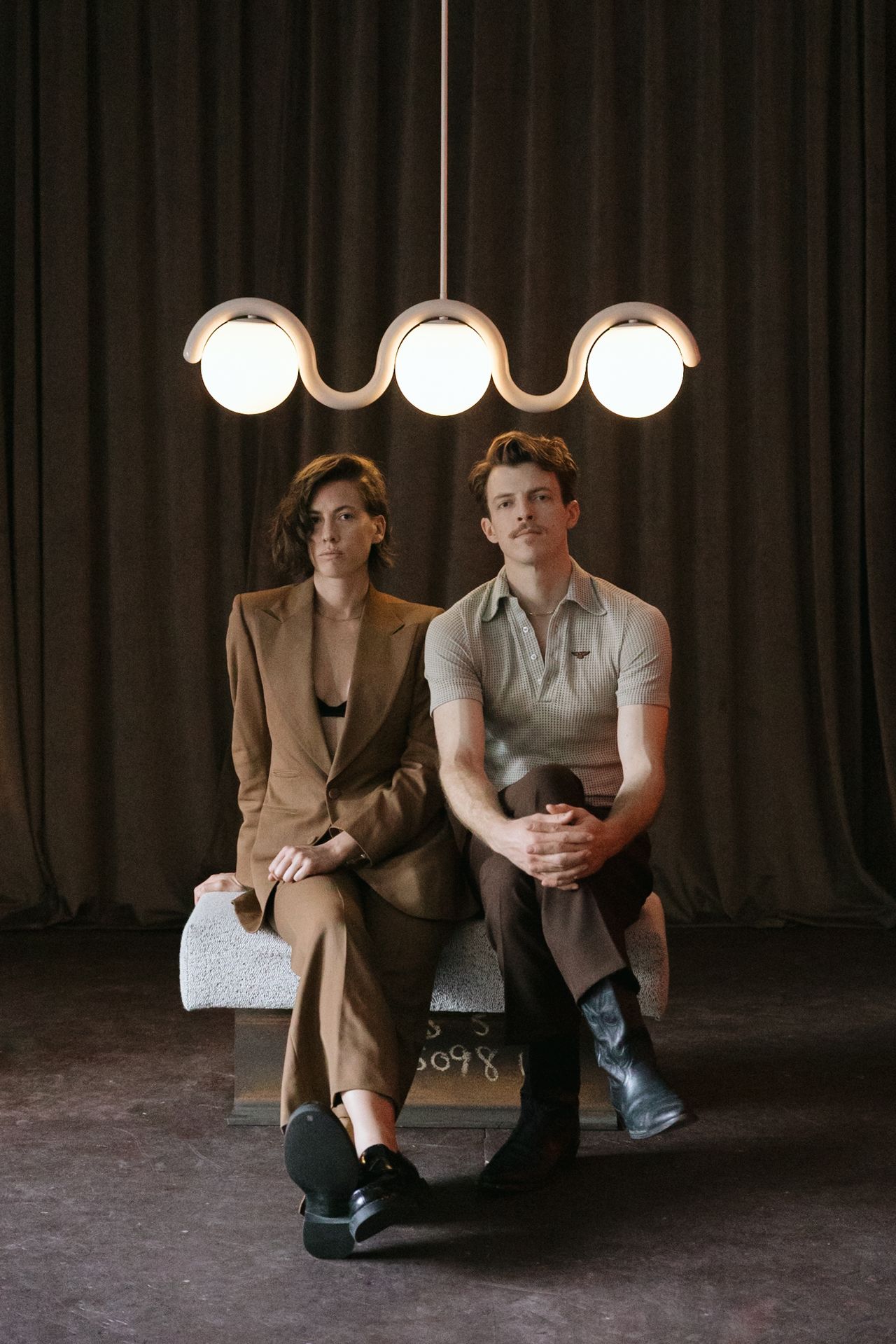A young couple dressed in matching brown-hued, smart casual outfits and black leather shoes sits on a bench under a whimsically shaped pendant chandelier composed of three spheres against a brown curtain.