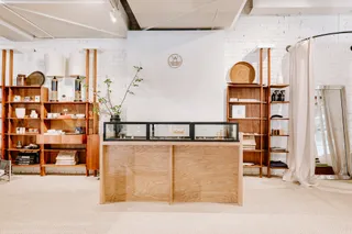 The organic interiors of a store feature wood, rattan, and linen furniture and accessories, including shelves, front desks, and other displaying units, on a beige carpet.