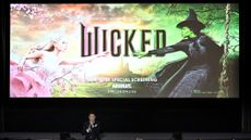 screening of Wicked movie, with the title still showing on a big screen and director Jon Chu speaking into a microphone