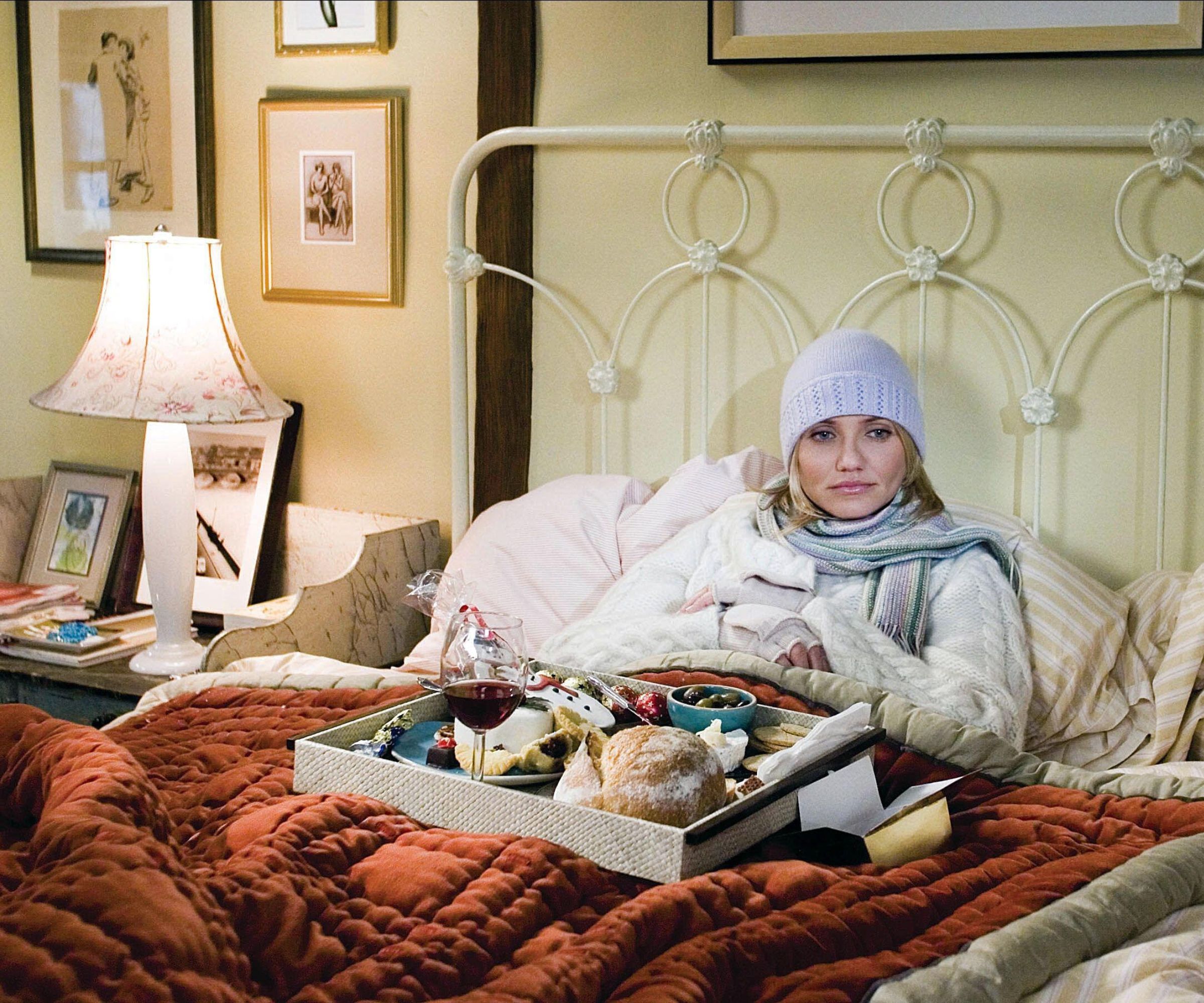 cameron diaz on set in the holiday movie sat in a wrought iron bed with striped beige bedding and a large rust colored quilt with a floral table lamp and marble washstand for a nightstand